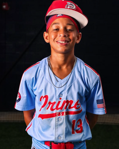 Young baseball player wearing a light blue jersey with red accents and a red cap, standing against a dark background.