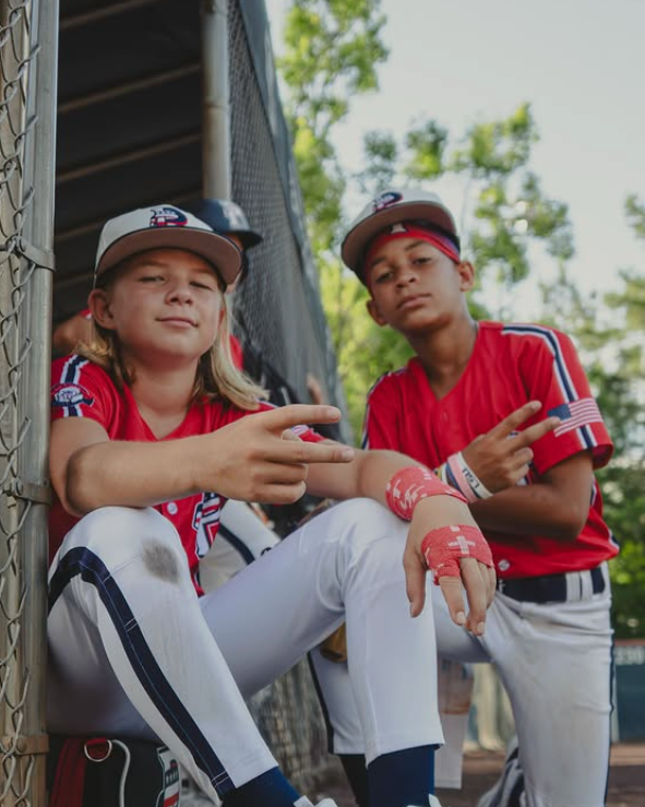 Two young baseball players in red and white uniforms sitting in a dugout.