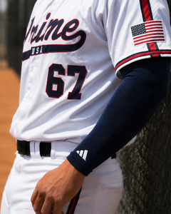 Baseball player wearing a white uniform with 'Prime USA' and number 67, leaning against a netted wall.