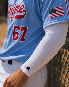 A close-up of a baseball player wearing a light blue jersey with red lettering and the number 67, layered with a white long-sleeve compression arm sleeve. The player is standing behind a chain-link fence on a baseball field.