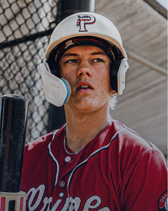 Baseball player in a red jersey and white helmet holding a bat, standing in front of a chain-link fence.