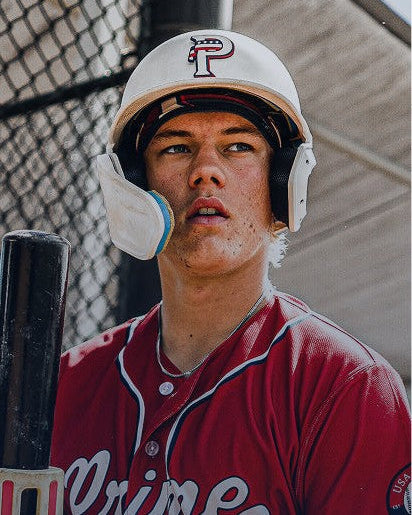 Baseball player in a red jersey and white helmet holding a bat, standing in front of a chain-link fence.