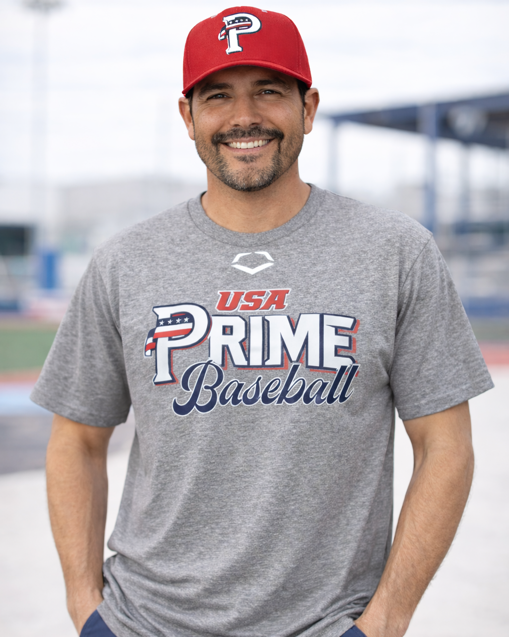 Man wearing a gray 'USA Prime Baseball' t-shirt and red cap at a baseball field.