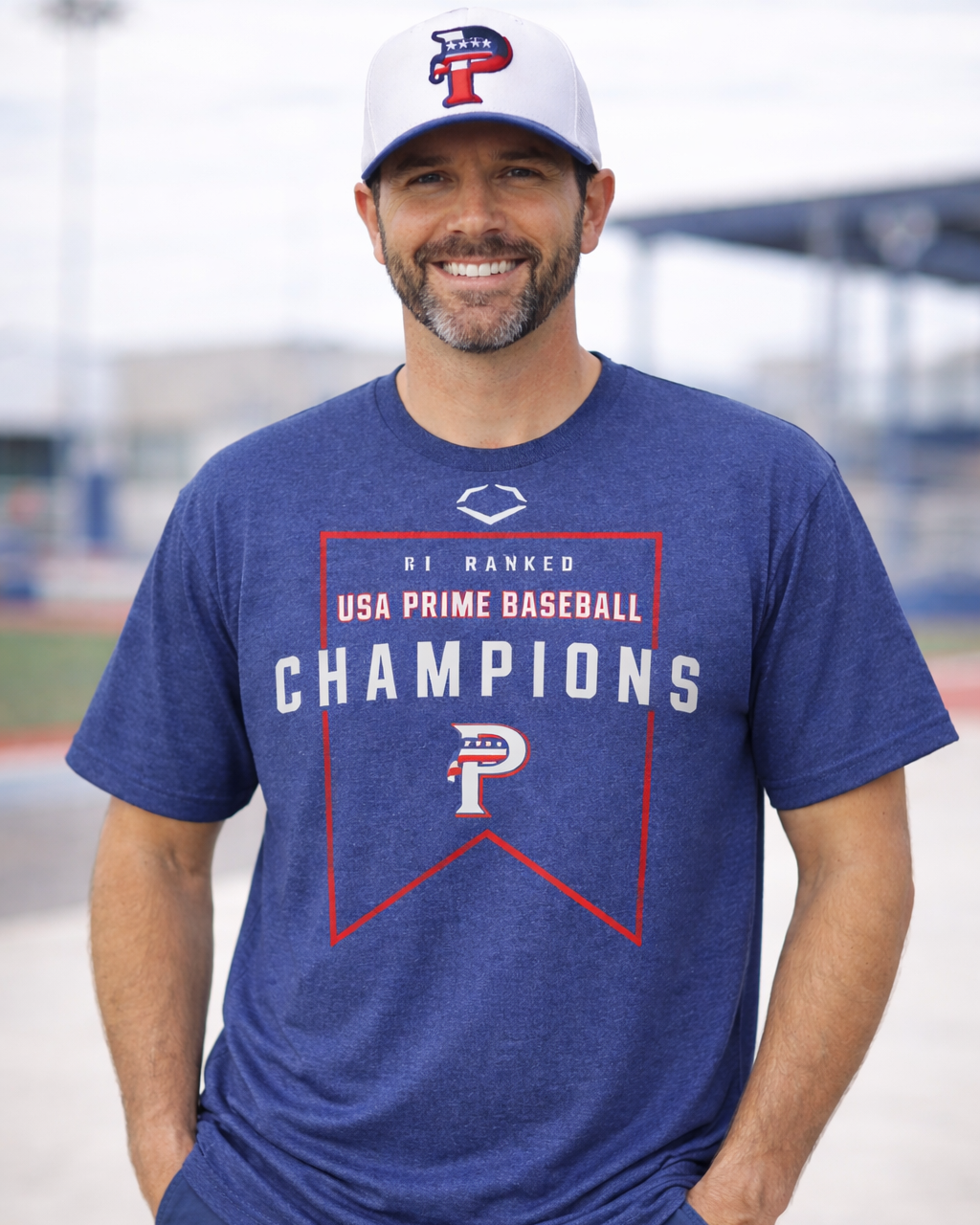 Man wearing a blue t-shirt with 'USA Prime Baseball Champions' text and logo, standing on a baseball field.